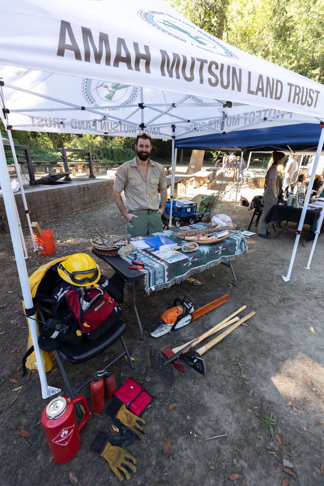 A man stands at the Amah Mutsun Land Trust booth, with hand tools, gloves, and a chainsaw laid out on the table.